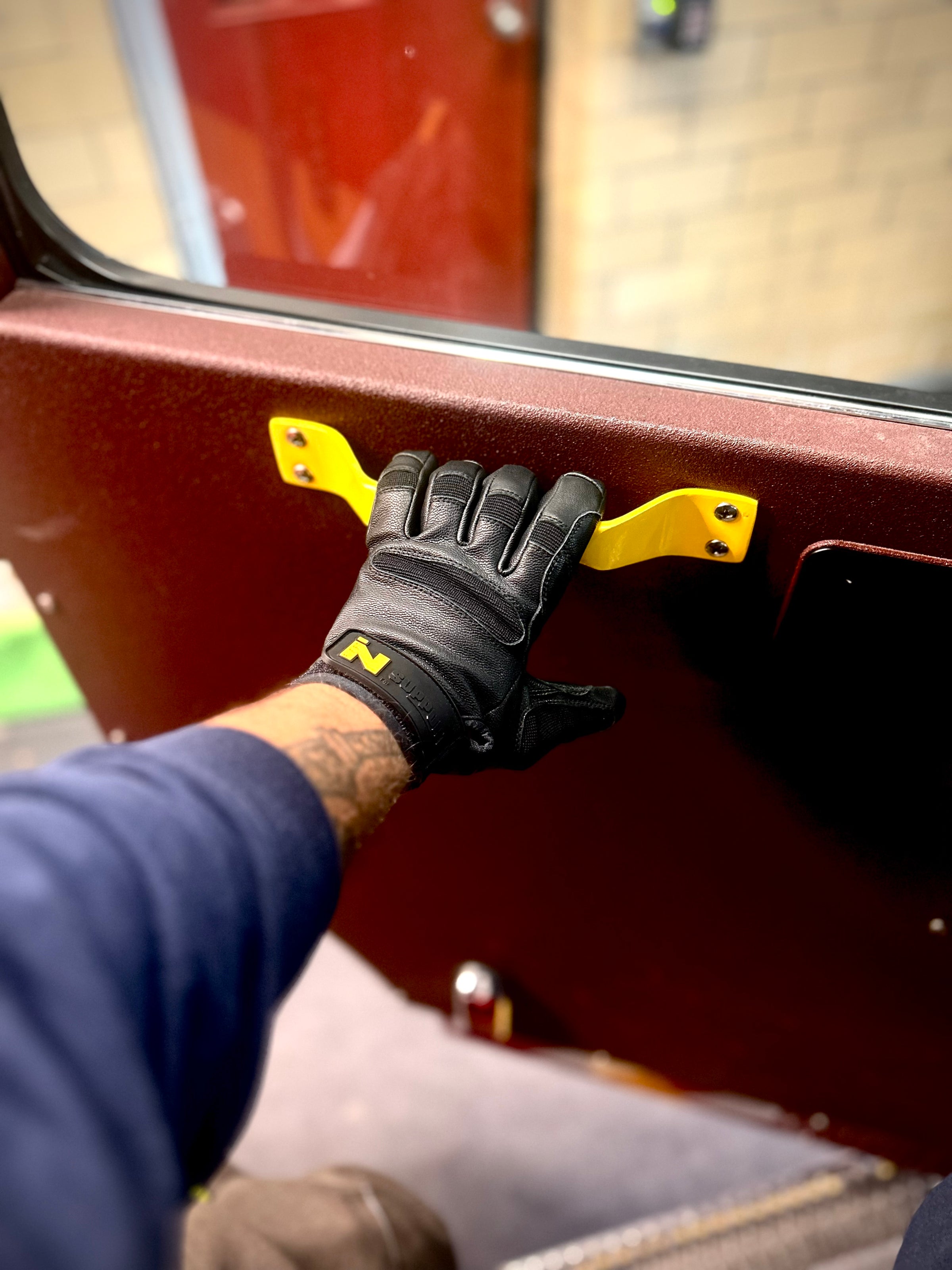 Firefighter wearing the FIRST IN - SUPPORT glove, holding a door handle to a fire truck.