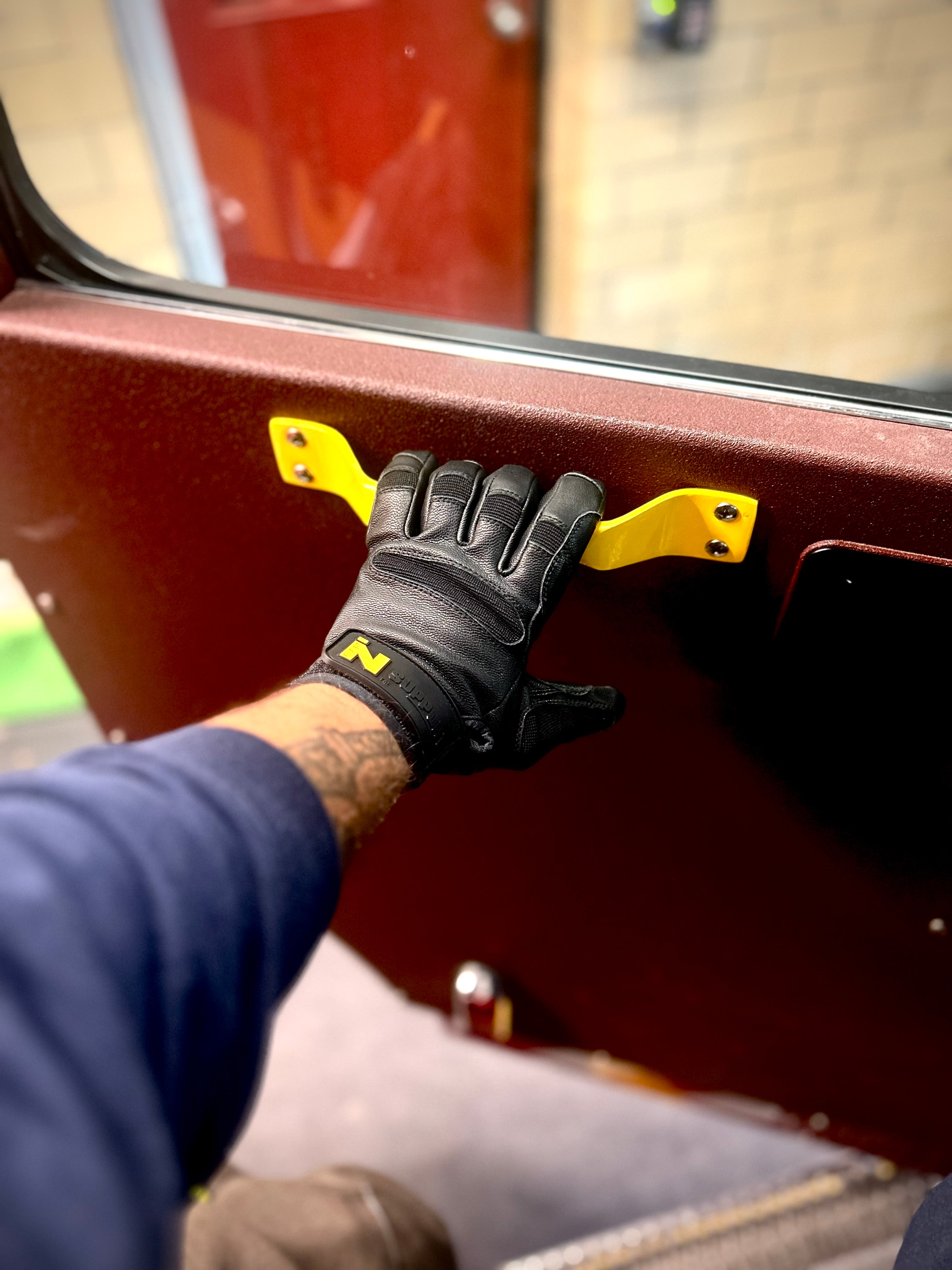 Firefighter wearing the FIRST IN - SUPPORT glove, holding a door handle to a fire truck.